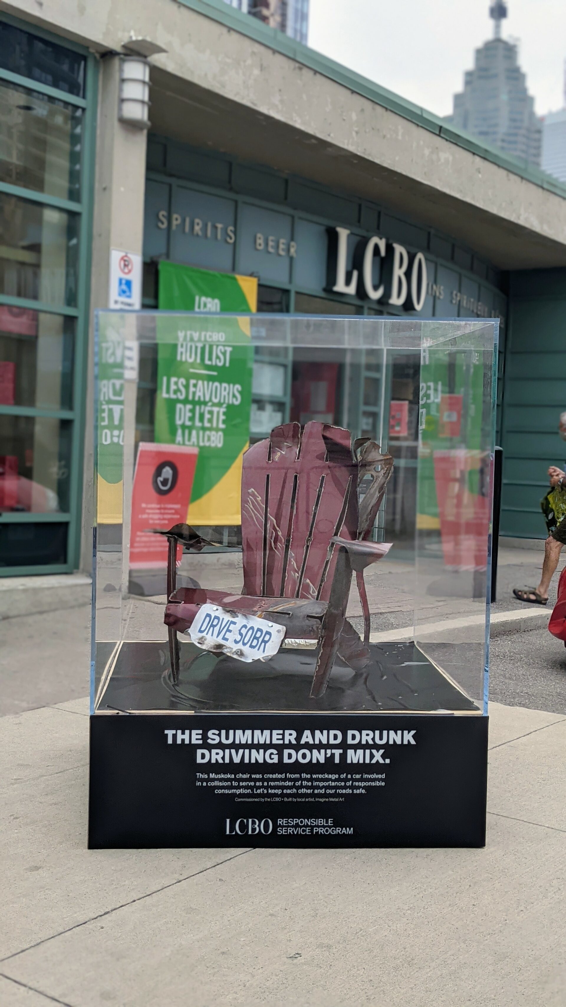 A red Muskoka chair, made entirely from reclaimed parts from car wrecks, sitting outside of an LCBO in Toronto. Text on the plaque reads: "The summers and drunk driving don't mix."