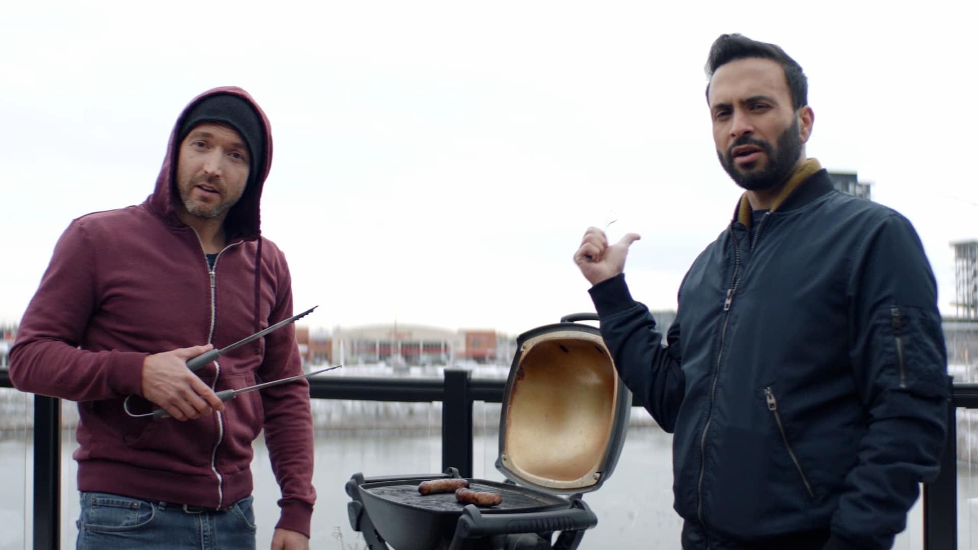 Two men standing over a BBQ, grilling sausages