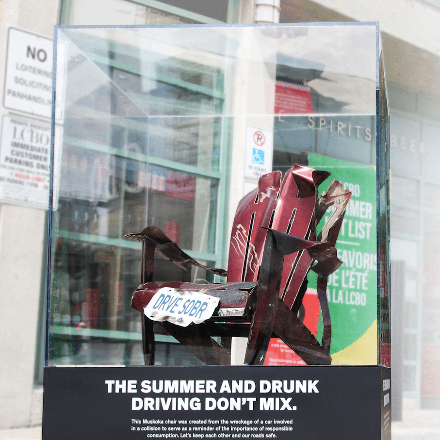 A red Muskoka chair, made entirely from reclaimed parts from car wrecks, sitting outside of an LCBO in Toronto. Text on the plaque reads: "The summers and drunk driving don't mix."
